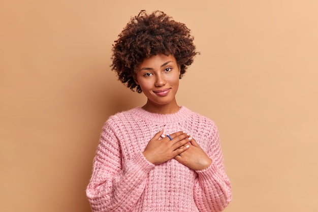 Beautiful curly haired woman shows her heartleft gratitude makes thankful gesture feels grateful for warm words appreciates your support and help wears knitted sweater isolated over brown wall