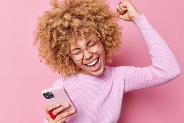 Beautiful curly haired young woman laughs positively shakes arm celebrates triumph uses modern smartphone cheers good news wears transparent eyeglasses casual jumper isolated over pink background