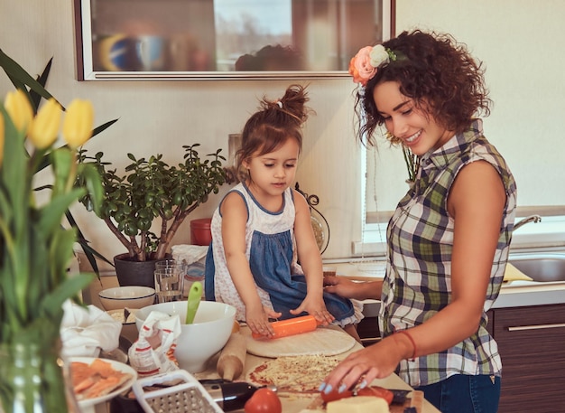 Beautiful curly Hispanic mother teaches her cute little daughter prepare pizza in the kitchen.