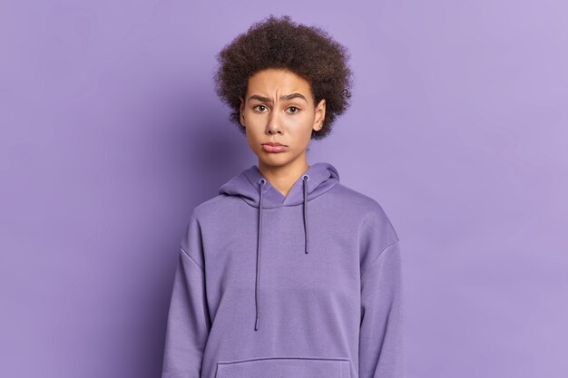 Beautiful curly sad teenage girl looks unhappily dressed in hoodie being offended at someone.