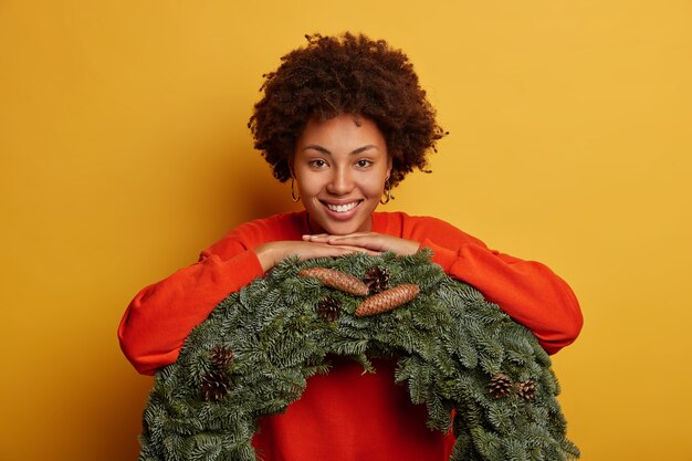 美しい巻き毛の女性は、緑の花輪に寄りかかって、カジュアルなジャンパーを着て、クリスマス前に家を飾り、歯を見せる笑顔を持ち、黄色の背景の上に孤立しています。