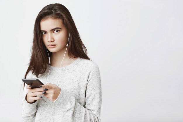beautiful cute european woman with long brown hair, holding smartphone while listening to music in headphones, expressing concern.