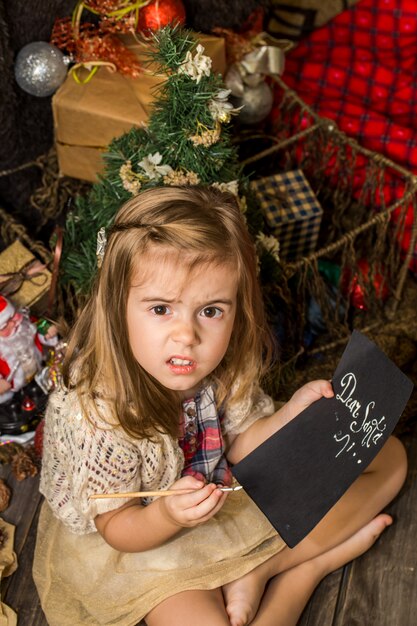 Beautiful cute little girl writes letter to Santa near Christmas decoration on the wooden floor