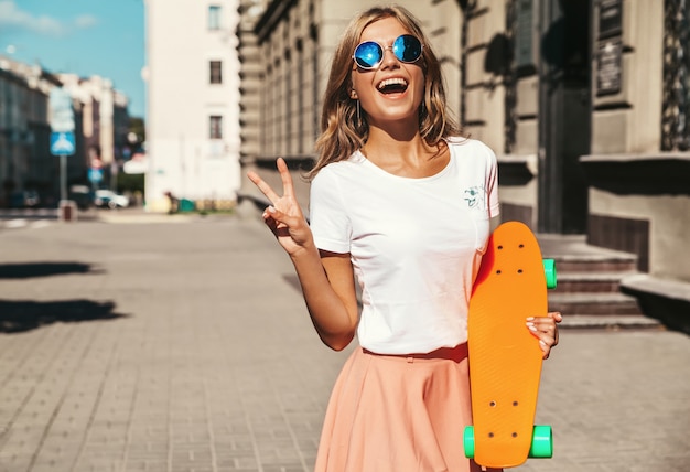 オレンジのペニースケートボードのポーズで夏の流行に敏感な服で美しいかわいい笑顔金髪ティーンエイジャーモデル。ピースサインを表示
