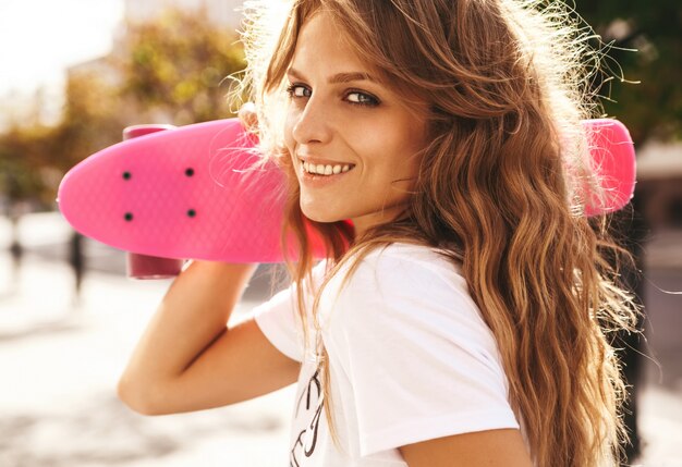 通りの背景にポーズをとってピンクのペニースケートボードと夏流行に敏感な白い服でメイクなしの美しいかわいい笑顔金髪ティーンエイジャーモデル