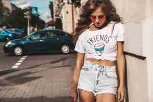 Free photo beautiful cute smiling blond teenager model without makeup in summer hipster white clothes with pink penny skateboard posing on the street background