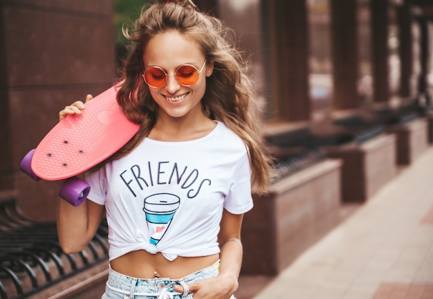 Beautiful cute smiling blond teenager model without makeup in summer hipster white clothes with pink penny skateboard posing on the street background