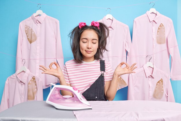 Beautiful dark haired Asian woman keeps eyes closed meditates during break after ironing dressed casually being busy housewife blue wall