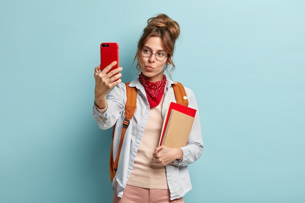 Beautiful dark haired Caucasian woman makes selfie portrait with cell phone, keeps lips folded, carries notebook and rucksack on back