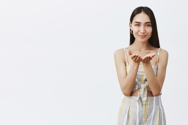 Beautiful dark-haired female with tanned skin in matching clothes holding palms near chest and smiling broadly while giving her heart and soul, standing over gray wall tender and gentle