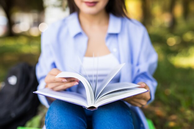 Beautiful dark-haired serious girl in jeans jacket and glasses reads book in park.