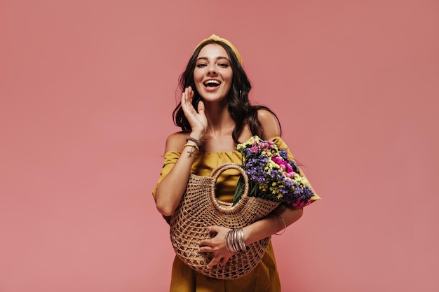 Beautiful dark haired woman in bright headband and yellow modern dress smiling and posing with cool straw handbag and colorful flowers