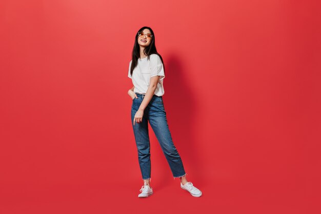 Beautiful dark-haired woman in loose jeans and white t-shirt posing on red wall