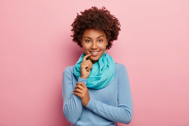 Beautiful dark haired woman talks lively, discusses something pleasant with friend, wears blue jumper and scarf, touches lip with index finger