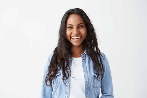 beautiful dark-skinned female with long brunette hair and broad happy smile wearing denim shirt enjoying good positive news concerning her promotion at work, posing isolated against white blank wall b