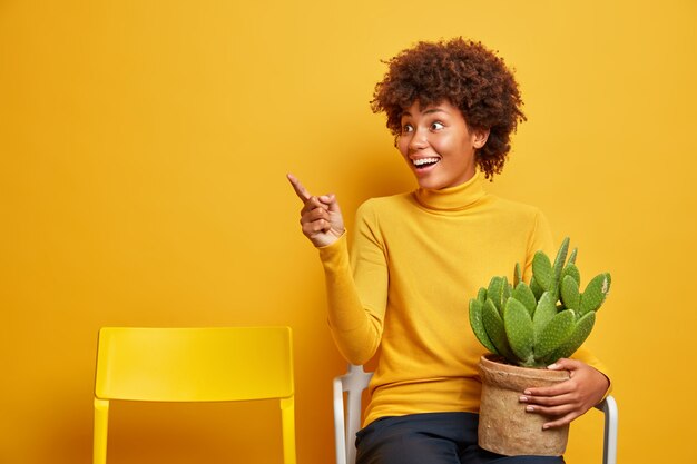 Beautiful dark skinned woman florist takes care of domestic plants carries pot of cactus poses on chair and points into distance with cheerful expression