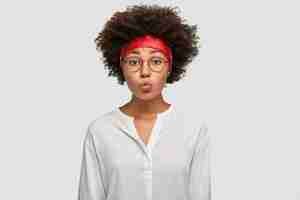 Free photo beautiful dark skinned woman pouts lips, has afro haircut, makes grimace, wears round spectacles, red headband and white shirt, stands against wall. facial expressions concept