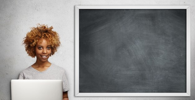 Beautiful dark-skinned young female teacher with stylish haircut checking papers of her students, using laptop in classroom