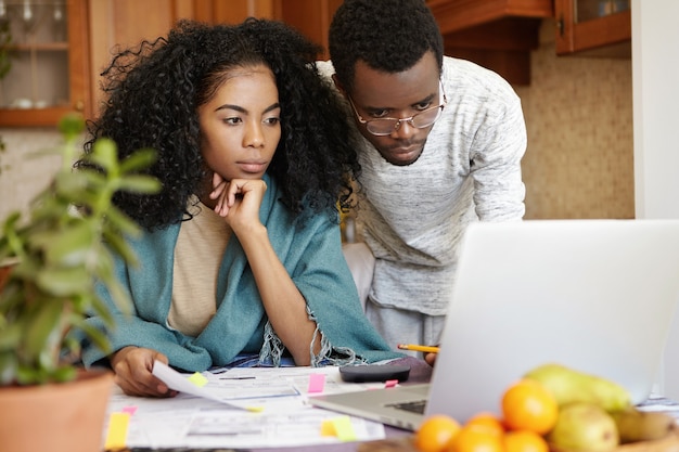 Bella giovane femmina dalla carnagione scura con acconciatura afro con sguardo preoccupato durante la gestione del bilancio familiare