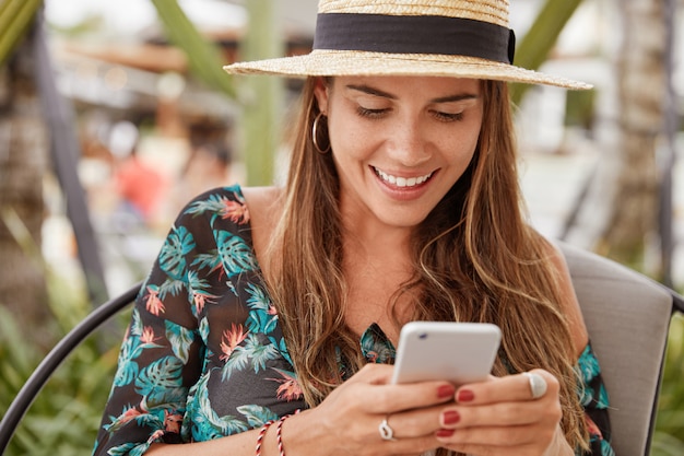 Beautiful delighted female tourist rests in exotic country, wears summer straw hat and blouse, types message on cell phone, connected to wireless internet at cozy cafeteria. People and vacation