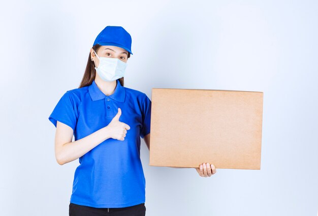 Beautiful deliverywoman in medical mask holding carton package and giving thumbs up. 