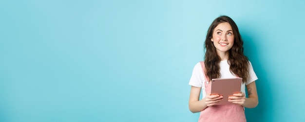 Free photo beautiful dreamy girl smiling holding tablet in hands and looking at upper left corner logo with tho