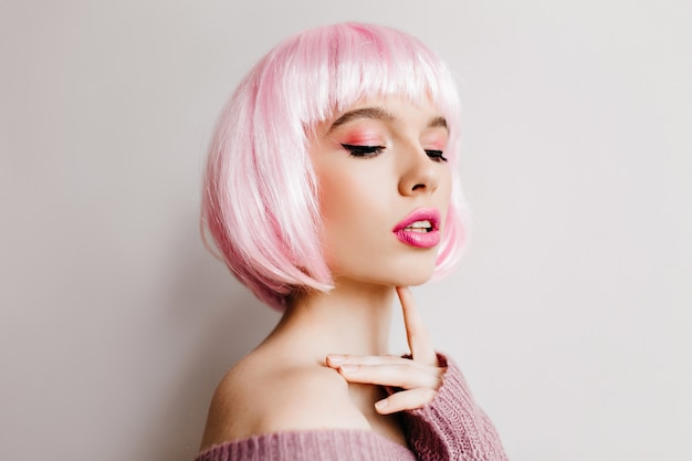 Красивая мечтательная женщина носит розовый peruke, позируя с закрытыми глазами. Фотография в помещении очаровательной девушки-модели с ярким макияжем в парике, стоящей на светлой стене.