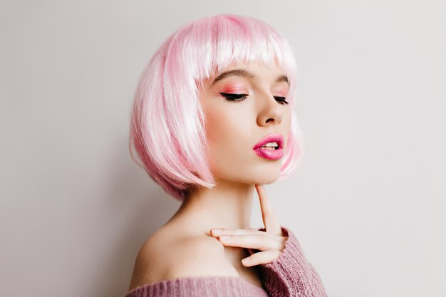 Beautiful dreamy woman wears pink peruke posing with eyes closed. Indoor photo of charming  female model with bright makeup in periwig standing on light wall.