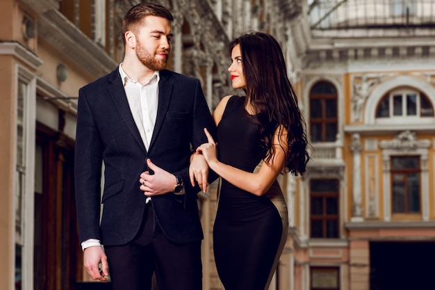 Beautiful elegant brunette with her husband  walking on the ale city streets . Enjoying there time, wearing black classic suit and long cocktail dress.