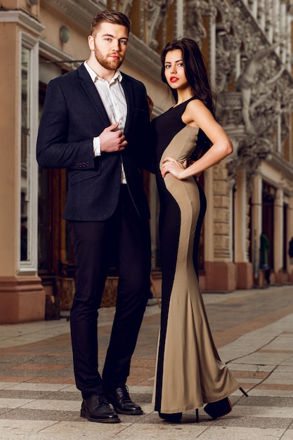 Beautiful elegant brunette with her husband  walking on the ale city streets . Enjoying there time, wearing black classic suit and long cocktail dress.