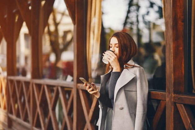 秋の街に立ってコーヒーを飲む美しいエレガントな女の子