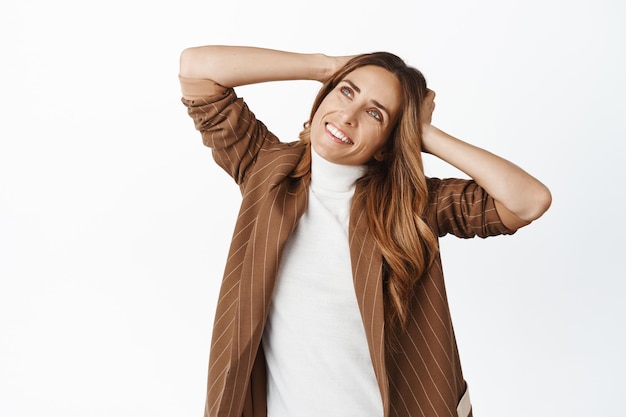 Beautiful and elegant middle aged woman smiling holding hands behind head and gazing up with dreamy pleased face expression white background