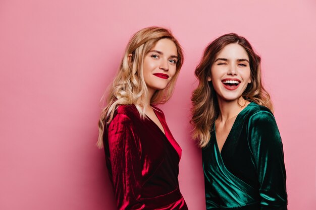 Beautiful elegant women standing on rosy wall. Enchanting blonde girl in red velvet dress posing with brunette sister.