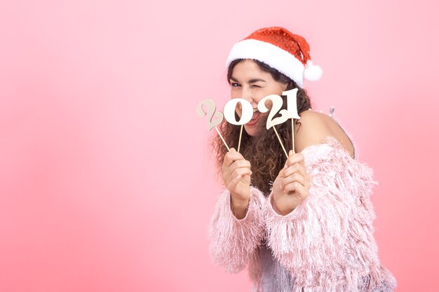 Красивая элегантная молодая брюнетка с вьющимися волосами в новогодней шапке на розовом студийном фоне позирует с деревянным номером для новогодней концепции