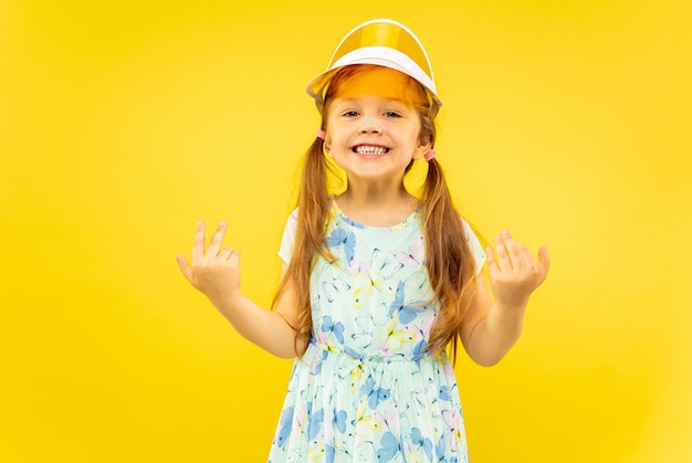 Бесплатное фото Красивая эмоциональная маленькая девочка, изолированные на желтом фоне. половинный портрет счастливого и празднующего ребенка в платье и оранжевой кепке. понятие лета, человеческих эмоций, детства.