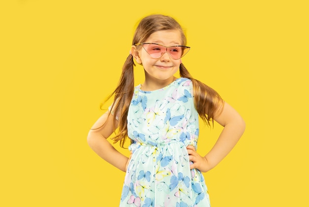 Beautiful emotional little girl isolated. portrait of happy child standing and wearing a dress and red sunglasses. Concept of summer, human emotions, childhood.