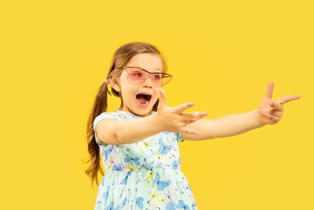 Beautiful emotional little girl isolated on yellow space. Half-lenght portrait of happy child standing and wearing a dress and red sunglasses