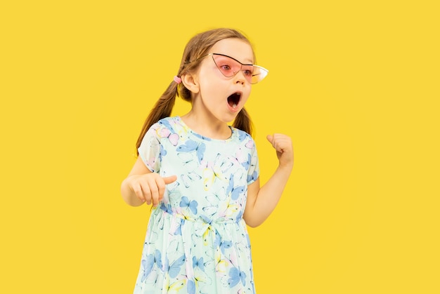 Beautiful emotional little girl isolated on yellow space. Half-lenght portrait of happy child standing and wearing a dress and red sunglasses