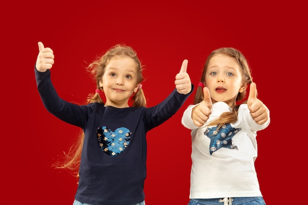 Beautiful emotional little girls isolated on red  space. Half-lenght portrait of happy sisters or friends pointing up