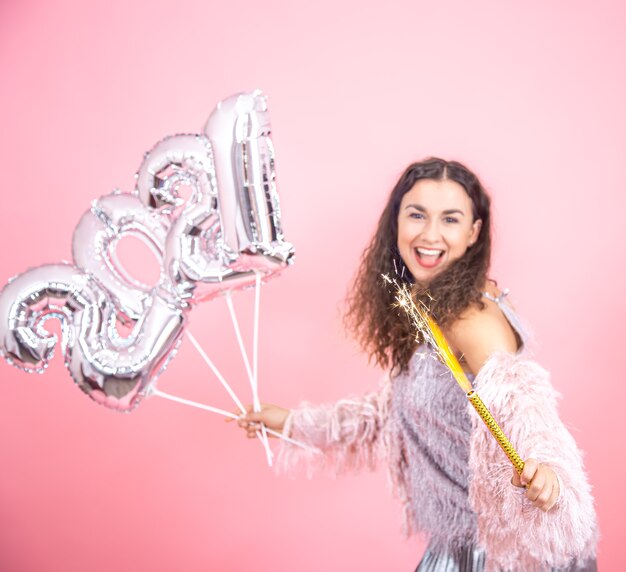 Beautiful emotional young brunette with curly hair festively dressed holding a fireworks candle in her hand and silver balloons for the new year concept
