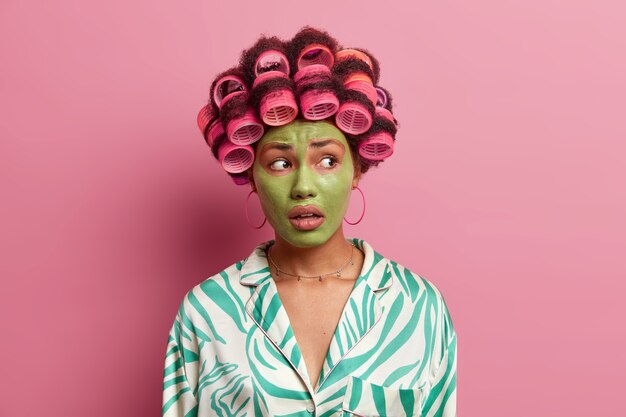 Beautiful ethnic woman has worried expression, looks away, applies green beauty mask for reducing fine lines, wears dressing gown isolated over pink . Cosmetology, wellness, hairstyling