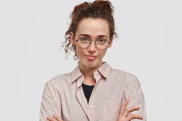 Beautiful European female feels bored to listen not interesting story from friend, keeps hands crossed, pretends listening attentively, has dark curly hair combed, wears round glasses and shirt