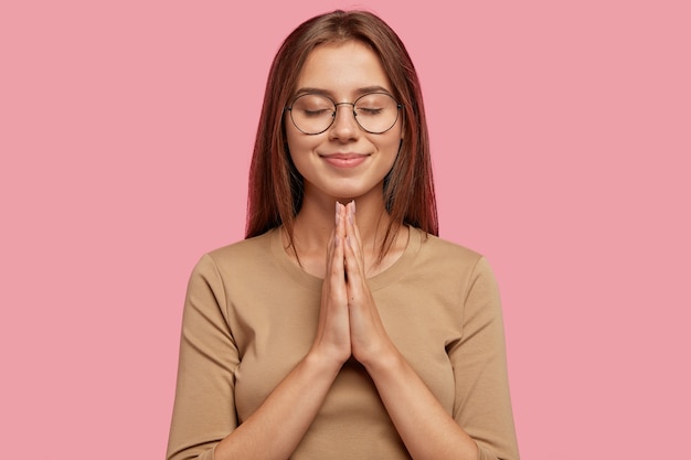 Бесплатное фото Красивая европейская женщина умоляет о помощи или просит извинений, пока держит руки в молитвенном жесте, носит круглые очки, темные волосы, одетая в повседневную одежду, изолирована на розовой стене