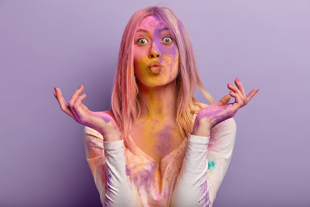 Beautiful European woman keeps lips folded, has surprised facial expression, spreads hands, wears casual clothes smeared with colored jumper, isolated over purple wall. Holi festival concept