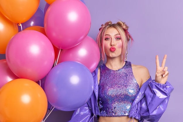 Beautiful European woman keeps lips folded makes peace gesture celebrates specical occasion poses with multicolored helium balloons