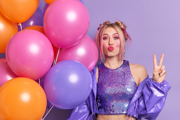 Beautiful European woman keeps lips folded makes peace gesture celebrates specical occasion poses with multicolored helium balloons