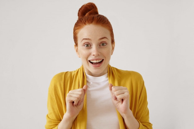 Free photo beautiful european woman with hair knot expressing her astonishment and excitement, clenching fists, raising eyebrows and looking with mouth opened.