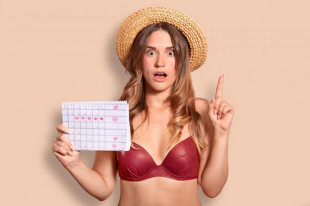 Beautiful European young female has surprised expression, raises index finger, dressed in red bikini and straw hat