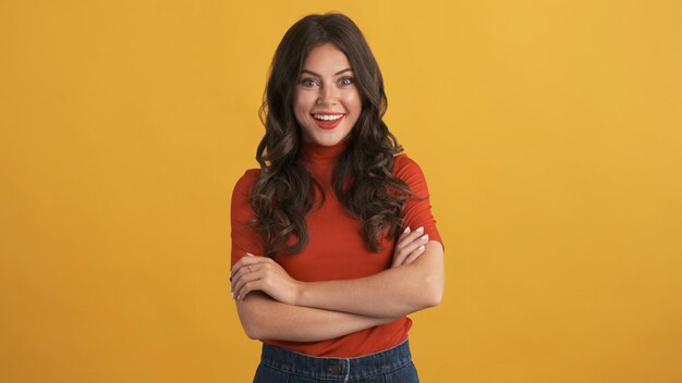 Beautiful excited brunette girl in red top with hands crossed happily looking in camera over colorful background