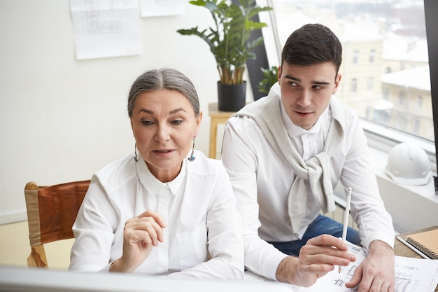 Beautiful experienced gray haired female architect and her skilled young male colleague enjoying common work, making drawings of new housing development project, sitting at desk and using computer